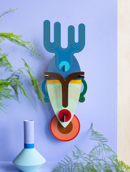 Bunte Karton Maske zum Aufhängen, Wandbehang, blau grün rot, fiktives Gesicht, auf blauer Wand mit Blumen 
